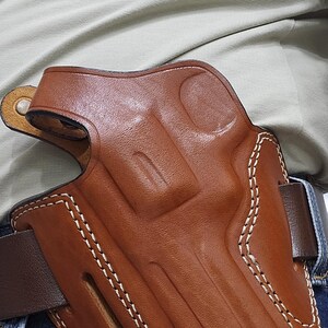 May include: A brown leather holster with white stitching, designed to be worn on a belt. The holster is a rich, medium brown color and appears to be made of high-quality leather. The stitching is clean and precise, adding to the holster's durability and aesthetic appeal.