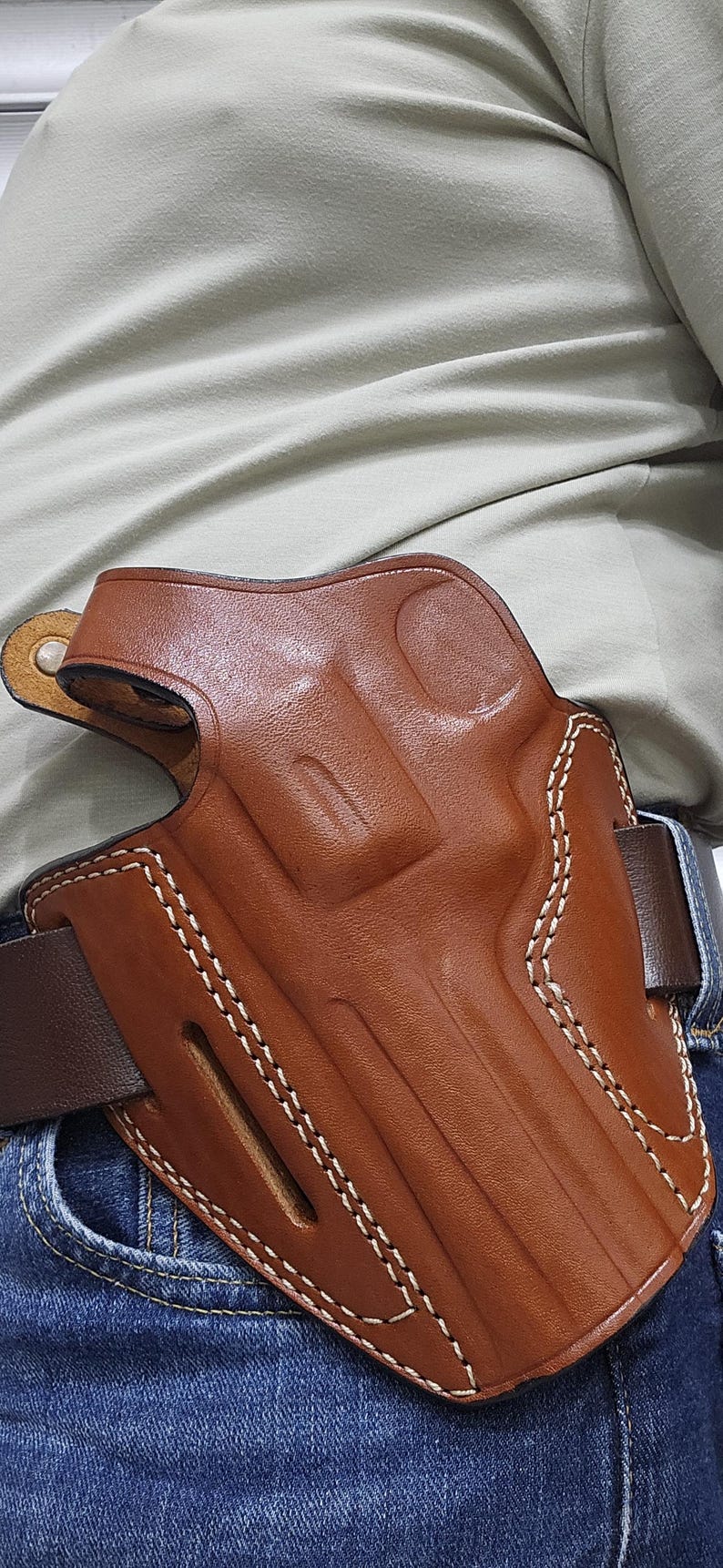 May include: A brown leather holster with white stitching, designed to be worn on a belt. The holster is a rich, medium brown color and appears to be made of high-quality leather. The stitching is clean and precise, adding to the holster's durability and aesthetic appeal.