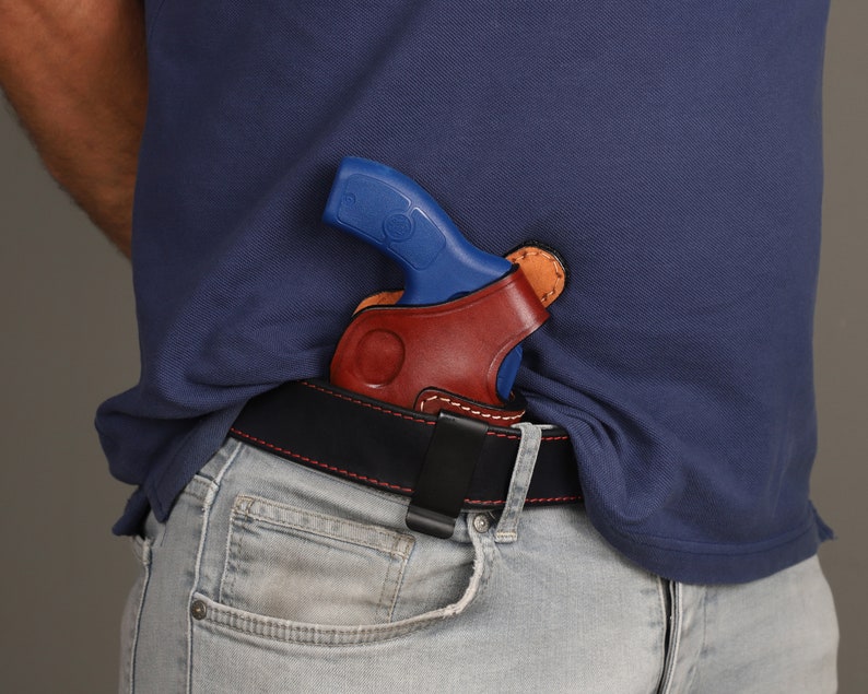 May include: A brown leather holster with a black belt clip, holding a blue and silver handgun. The holster is worn on a person's waist.