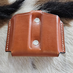 Może przedstawiać: Brązowa skórzana kabura z dwoma srebrnymi nitami. Kabura jest obszyta białą nicią wzdłuż krawędzi. Tło to dywan z krowiej skóry z brązowym, czarnym i białym futrem.