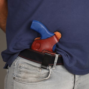 Puede incluir: Una funda de cuero marr&oacute;n con un clip de cintur&oacute;n negro, que contiene una pistola azul y plateada. La funda se lleva en la cintura.