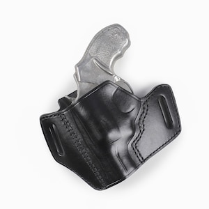 Può includere: Una fondina in pelle nera con una pistola argentata all'interno. La fondina è realizzata in pelle liscia e lucida con cuciture dettagliate. L'impugnatura della pistola è visibile, mostrando una superficie strutturata.