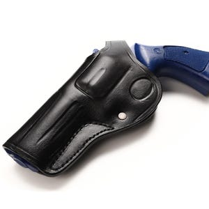 Può includere: Una fondina in pelle nera con una pistola blu. La fondina è realizzata in pelle liscia e lucida e presenta cuciture dettagliate. La pistola ha un'impugnatura blu e una canna nera. Gli oggetti sono su uno sfondo bianco.