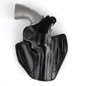 Può includere: Un fodero in pelle nera per un revolver. Il fodero ha una patta che copre la guardia del grilletto e un passante per la cintura sul retro.