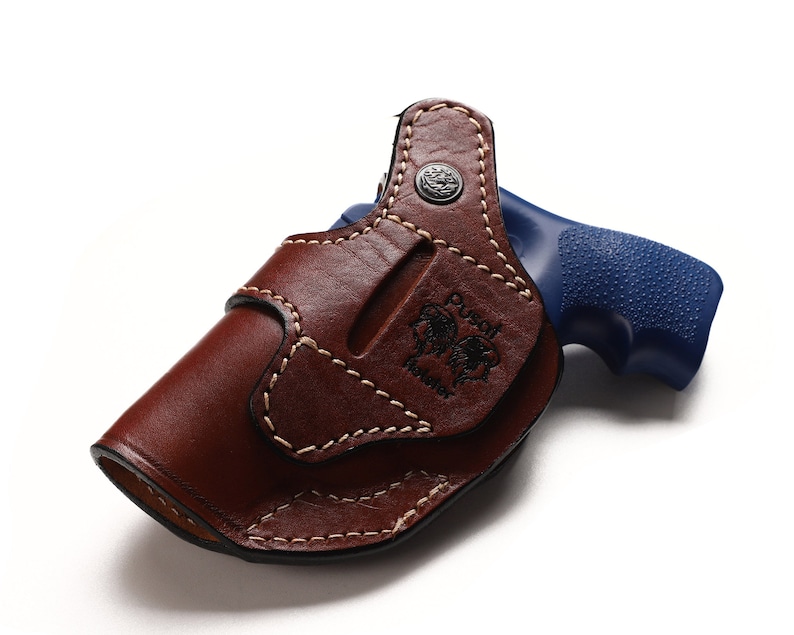 May include: A brown leather holster with a stitched design and the text "Dust Devil Holster" embossed on the side. A blue handgun is partially visible in the holster.