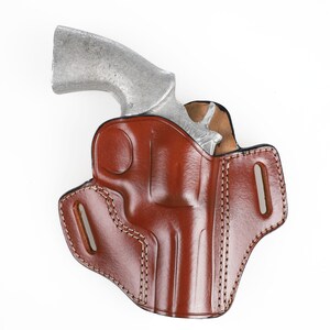 Peut inclure: Un étui en cuir marron avec un revolver argenté à l'intérieur. L'étui est cousu sur les bords et comporte deux passants de ceinture.