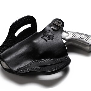 Puede incluir: Funda de cuero negra con un rev&oacute;lver plateado en su interior. La funda tiene un bucle para el cintur&oacute;n y un cierre a presi&oacute;n. El rev&oacute;lver tiene un acabado plateado y una empu&ntilde;adura negra.