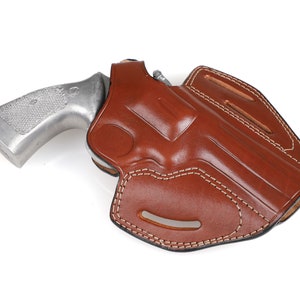 May include: A brown leather holster with white stitching, designed to hold a handgun. The grip of a silver-colored revolver is partially visible, resting inside the holster. The holster has a molded shape and a smooth, polished finish.