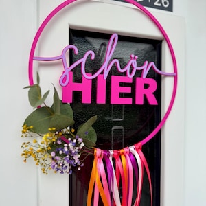 Puede incluir: Una vibrante corona de puerta circular rosa con las palabras alemanas "Schön HIER" en un diseño en capas. La corona está adornada con cintas de colores y un pequeño arreglo floral. La corona está colgada en una puerta blanca.