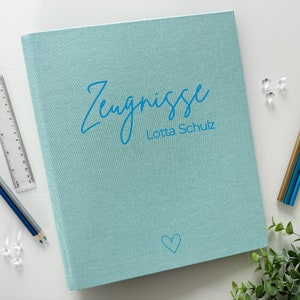 Könnte beinhalten: Ein hellblauer Ordner mit dem Text "Zeugnisse" und "Lotta Schulz" in blauer Schrift. In der unteren rechten Ecke befindet sich ein kleines blaues Herz.