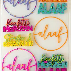Puede incluir: Varios letreros decorativos coloridos con la palabra "Alaaf" en varios estilos, incluyendo un dise&ntilde;o circular y letras en bloque. Otros letreros presentan frases como "Konfetti im Herzen" y nombres. Los letreros son de colores brillantes.
