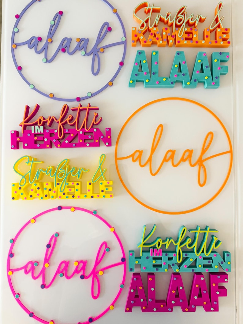 Puede incluir: Varios letreros decorativos coloridos con la palabra "Alaaf" en varios estilos, incluyendo un dise&ntilde;o circular y letras en bloque. Otros letreros presentan frases como "Konfetti im Herzen" y nombres. Los letreros son de colores brillantes.