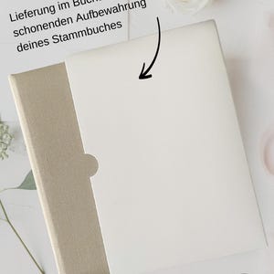 May include: A white book with a beige slipcover. The book is closed and the slipcover is folded over the top. The text on the slipcover reads "Lieferung im Buchschuber zur schonenden Aufbewahrung deines Stammbuches".