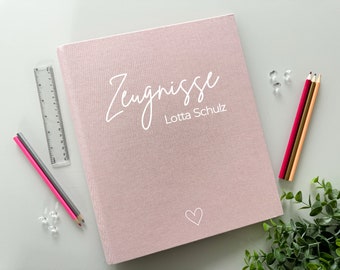 以下が含まれることがあります： 白い文字で「Zeugnisse」と「Lotta Schulz」と書かれたピンクの布製バインダー。右下隅に小さな白いハートがある。