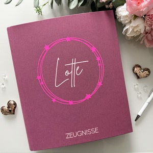 Könnte beinhalten: Ein rosa Ordner mit einem weißen Kreis und dem Namen "Lotte" in Kursivschrift. Der Kreis ist mit rosa Sternen umrandet. Der Ordner trägt außerdem das Wort "ZEUGNISSE" in weiß unten.