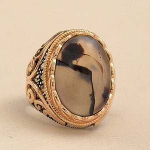Puede incluir: Un anillo de plata con un engaste ovalado de tono dorado que presenta una piedra de ágata marrón y blanca. El anillo tiene un diseño detallado y ornamentado.