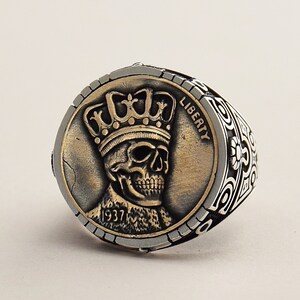 May include: A silver ring with a detailed design featuring a skull wearing a crown. The skull is on a coin-shaped piece with the word "LIBERTY" and the year "1937" inscribed.