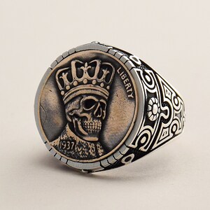 May include: A silver ring with a detailed design featuring a skull wearing a crown. The skull is on a coin-shaped piece with the words "LIBERTY" and "1937" visible.