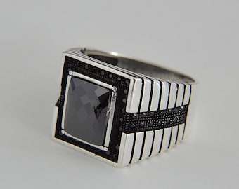 May include: A silver ring with a black square stone set in a silver frame. The ring has a band with a series of silver and black lines.