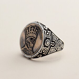 May include: A silver ring with a round, detailed design. The center of the ring features a skull wearing a crown, with the words "LIBERTY" and "1937" visible. The band of the ring is decorated with intricate geometric patterns.