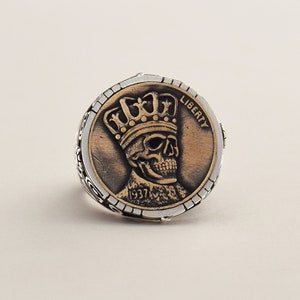 May include: A silver ring with a copper-colored coin design featuring a skull wearing a crown. The coin reads "LIBERTY" and "1937".