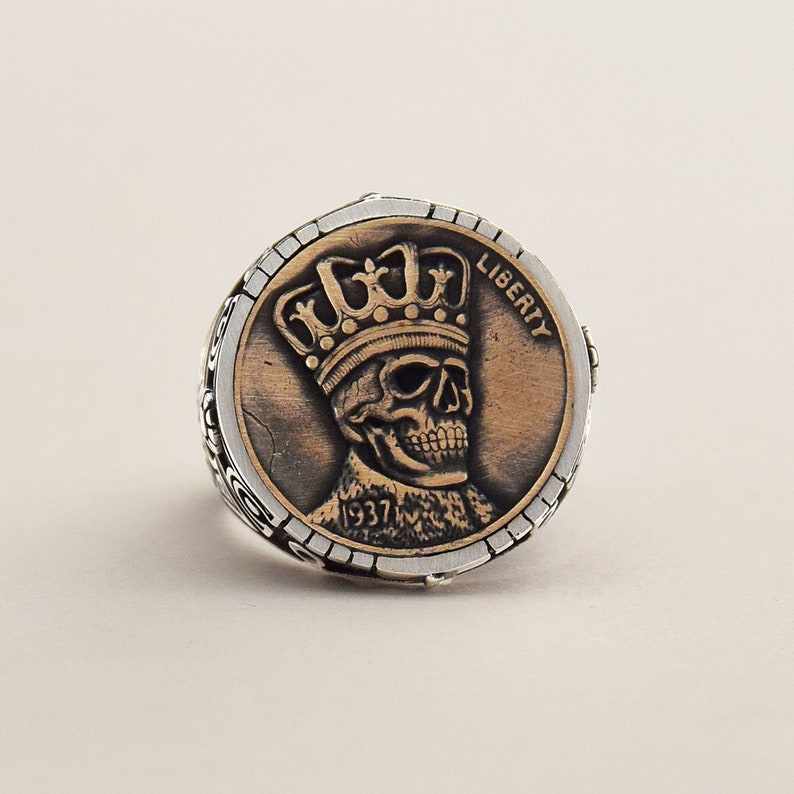 May include: A silver ring with a copper-colored coin design featuring a skull wearing a crown. The coin reads "LIBERTY" and "1937".