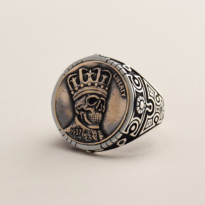May include: A silver ring with a detailed design featuring a skull wearing a crown. The skull is on a coin-shaped piece with the words "LIBERTY" and "1937" visible.
