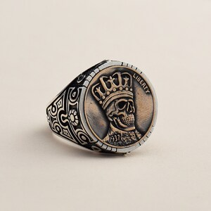 May include: A silver ring with a black and silver patterned band. The ring features a copper-colored coin with a skull wearing a crown and the word "LIBERTY" inscribed on the coin. The coin also has the year "1937" inscribed on it.