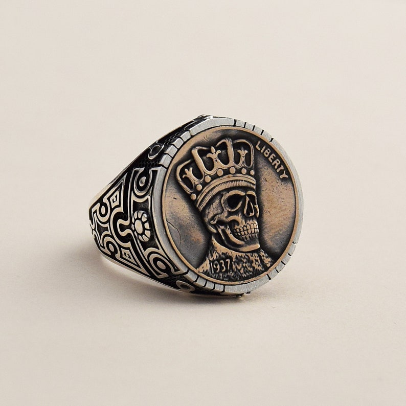May include: A silver ring with a black and silver patterned band. The ring features a copper-colored coin with a skull wearing a crown and the word "LIBERTY" inscribed on the coin. The coin also has the year "1937" inscribed on it.
