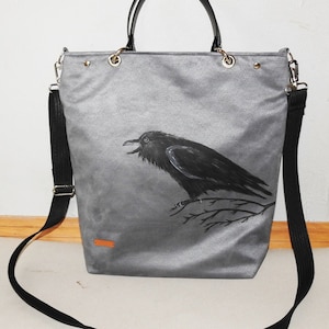 May include: A gray canvas tote bag with a black raven illustration. The bag has black shoulder straps and black handles.