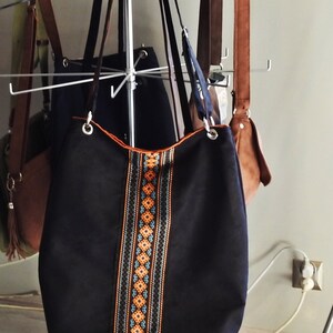 May include: A black tote bag with orange, blue, and black geometric patterned trim. The bag is hanging on a metal rack with other bags.