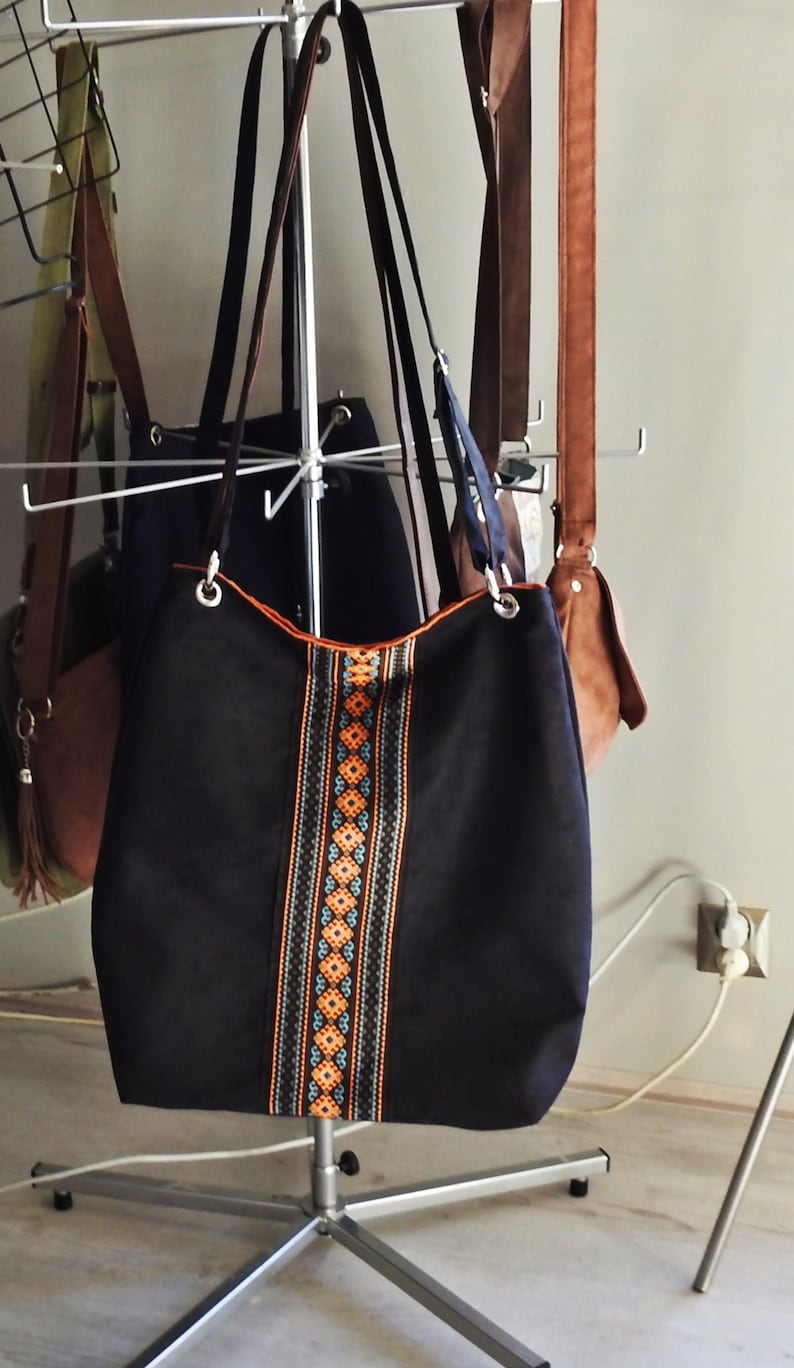 May include: A black tote bag with orange, blue, and black geometric patterned trim. The bag is hanging on a metal rack with other bags.