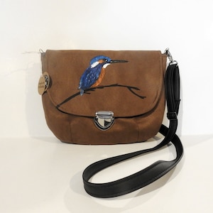 May include: A brown suede crossbody bag with a painted image of a blue kingfisher perched on a branch. The bag has a silver clasp and a black adjustable strap.