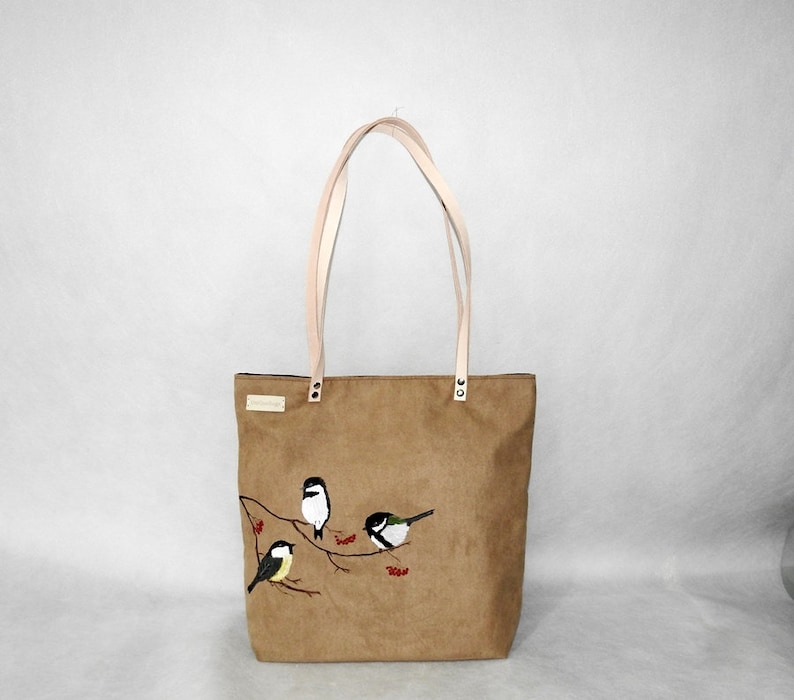May include: A brown tote bag with a light brown fabric and white straps. The bag has an embroidered design of four birds on a branch with red berries.