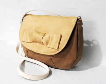 May include: A tan and brown suede crossbody bag with a large bow on the front flap. The bag has a white strap.