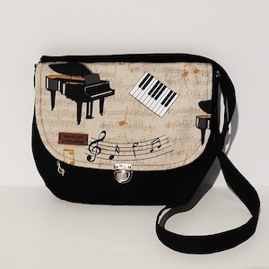 Può includere: Borsa a tracolla nera e beige a tema musicale. La borsa presenta un pianoforte nero, note musicali e una tastiera. La borsa ha una tracolla nera e una chiusura argentata.