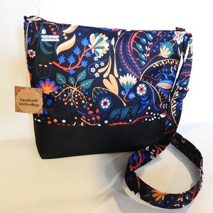 Può includere: Una borsa a tracolla nera con una stampa floreale che presenta fiori e foglie colorati su uno sfondo nero. La borsa ha una tracolla nera e un'etichetta marrone con la scritta "handicraft UniQueBags".