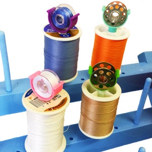 May include: A blue plastic thread rack with four spools of thread. The spools are white, orange, beige, and blue. Each spool has a plastic thread guide with a small metal ring.