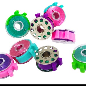 May include: Assortment of colorful plastic bobbins for sewing machines. The bobbins are in various colors, including green, pink, purple, and blue. Each bobbin has a clear plastic spool and a metal center.