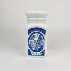May include: White ceramic tea bag container with a lid. The container features a blue and white design with a circular illustration and the words "TEA BAGS" at the bottom. The container is rectangular with rounded edges.