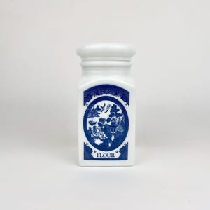 May include: White ceramic flour container with a lid. The container features a blue and white design with a landscape scene and the word "FLOUR" at the bottom. The container is rectangular with rounded edges.