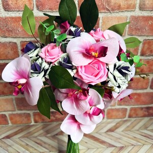 A hand tied bouquet made with pink orchids, book page roses layered with purple cardstock, pink foam roses, faux eucalyptus picks and fern greens. The bouquet has exposed stems and is tied with a matching purple ribbon.