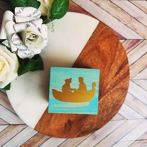 A wooden ring box or a wooden jewelry box stained in a vivid aqua color, with gold vinyl spelling showing the couple during their first moment together.