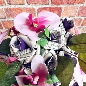 A hand tied bouquet made with pink orchids, book page roses layered with purple cardstock, pink foam roses, faux eucalyptus picks and fern greens. The bouquet has exposed stems and is tied with a matching purple ribbon.