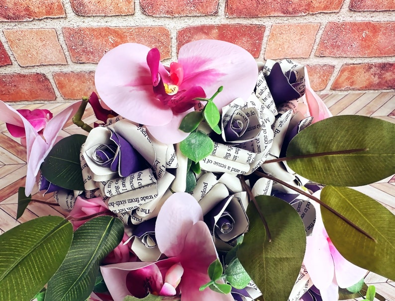 A hand tied bouquet made with pink orchids, book page roses layered with purple cardstock, pink foam roses, faux eucalyptus picks and fern greens. The bouquet has exposed stems and is tied with a matching purple ribbon.