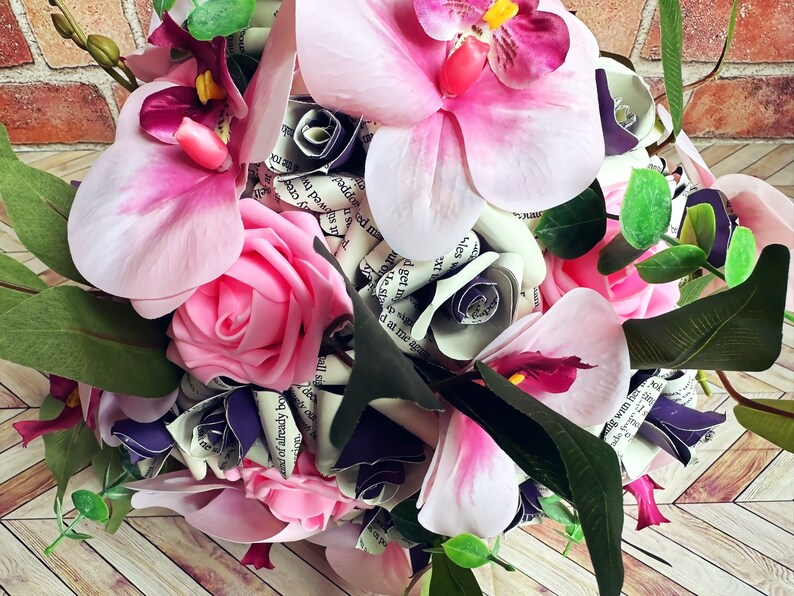 A hand tied bouquet made with pink orchids, book page roses layered with purple cardstock, pink foam roses, faux eucalyptus picks and fern greens. The bouquet has exposed stems and is tied with a matching purple ribbon.