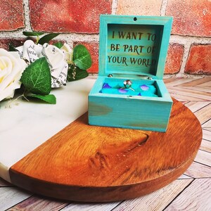 A wooden ring box or a wooden jewelry box stained in aqua, with gold vinyl saying I want to be part of your world. A cushion is wrapped in mermaid scale fabric where a ring or necklace can sit. There are built in barrel hinges and a magnetic latch.