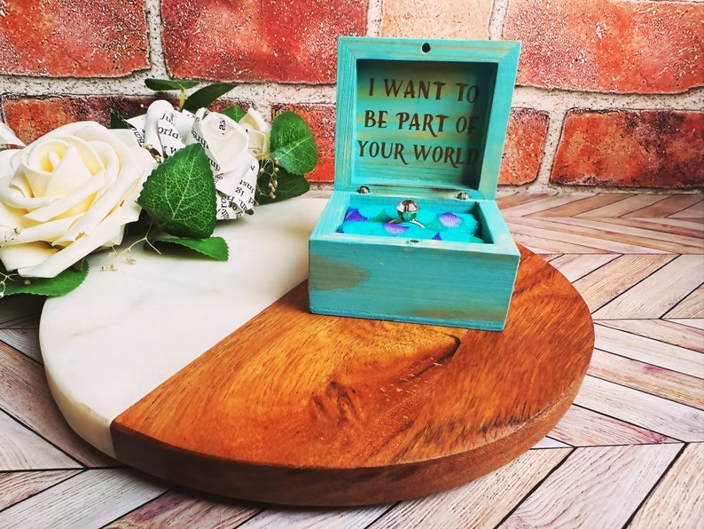 A wooden ring box or a wooden jewelry box stained in aqua, with gold vinyl saying I want to be part of your world. A cushion is wrapped in mermaid scale fabric where a ring or necklace can sit. There are built in barrel hinges and a magnetic latch.