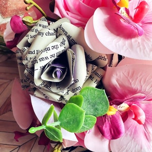 A hand tied bouquet made with pink orchids, book page roses layered with purple cardstock, pink foam roses, faux eucalyptus picks and fern greens. The bouquet has exposed stems and is tied with a matching purple ribbon.