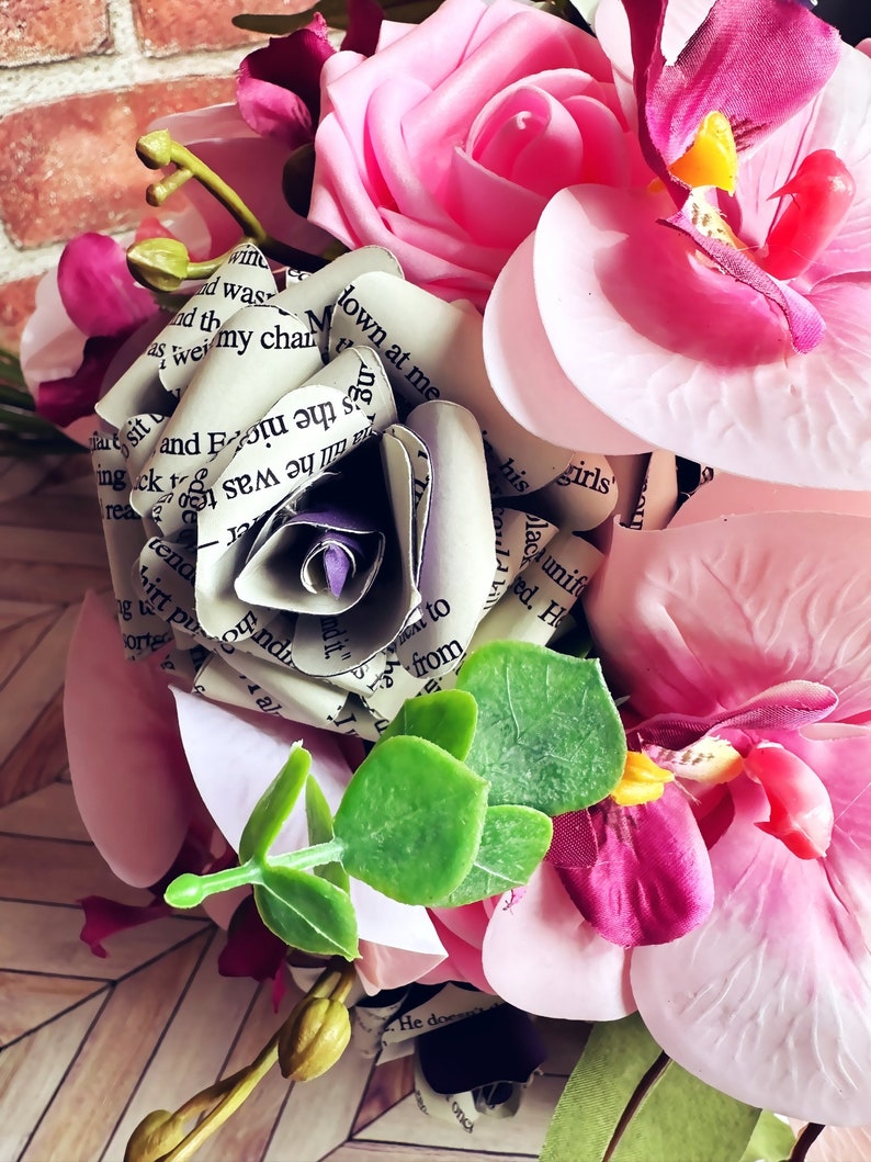 A hand tied bouquet made with pink orchids, book page roses layered with purple cardstock, pink foam roses, faux eucalyptus picks and fern greens. The bouquet has exposed stems and is tied with a matching purple ribbon.